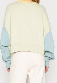 Persoon in een losse, gekleurde sweater met een crèmekleurig lichaam en lichtblauwe mouwen, gecombineerd met hoge witte broek, gezien vanaf de achterkant.