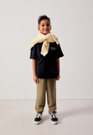 Nasmejan fant, oblečen v črno majico z napisom "American Vintage", bež hlače in črne superge, z svetlo rumeno jopico, premetano čez ramena.