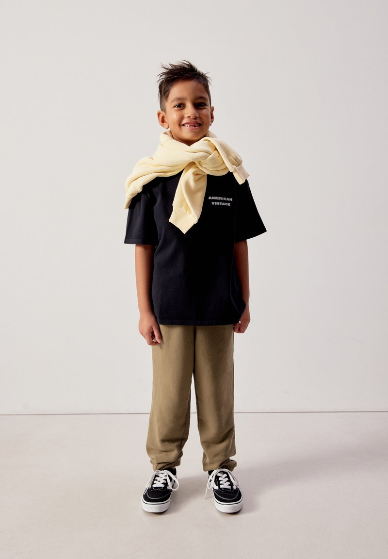 Naeratav poiss, kes kannab musta „American Vintage“ T-särki, beežide pükse, musti tossusid ning kelle õlgadele on heledat kollast pusa visatud.