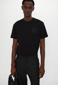 Camiseta de algodón negra con mangas cortas y cuello redondo. Presenta un logo tonal en el pecho, combinada con pantalones oscuros y un bolso.