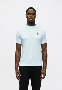 Αχάριστο μπλε κοντομάνικο μπλουζάκι από απαλό ύφασμα, με ένα μικρό ασπρόμαυρο λογότυπο στην αριστερή πλευρά του στήθους.