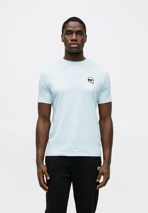 Helles blaues kurzärmeliges T-Shirt aus einem weichen Stoff, mit einem kleinen schwarz-weißen grafischen Logo im linken Brustbereich.