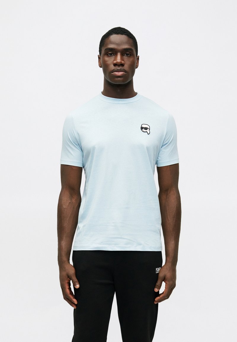 Αχάριστο μπλε κοντομάνικο μπλουζάκι από απαλό ύφασμα, με ένα μικρό ασπρόμαυρο λογότυπο στην αριστερή πλευρά του στήθους.