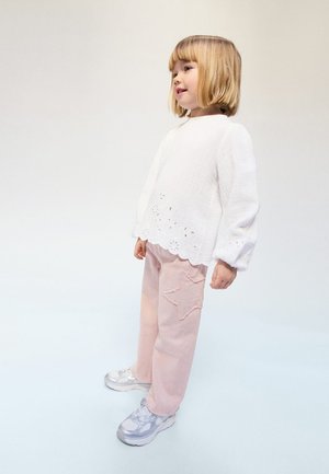 Mlajši otrok s svetlim bobi odbitkom, oblečen v belo majico z dolgimi rokavi in svetlo rožnate hlače z zvezdico, ki stoji na enobarvnem svetlem ozadju.