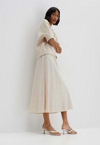 Pull en tricot crème à manches courtes, associé à une jupe midi fluide beige clair et des sandales à talons argentés, se tenant contre un fond blanc.