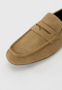 Mocasin de gamuza marrón con una capellada de puntera cosida, tira de cuero decorativa y suela flexible. Textura suave, diseño minimalista.
