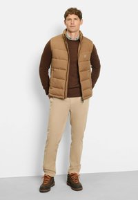 Braune Weste in Puffer-Optik über einem braunen Pullover, beigen Hosen und braunen Stiefeln mit orangefarbenen Schnürsenkeln. Die Weste verfügt über einen Reißverschluss und ein Logo-Detail.