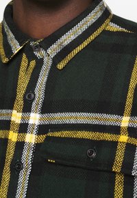 Primo piano di una camicia a quadri verde scuro con strisce gialle, bianche, bottoni neri e una tasca sul petto con bottone, su una pelle di tono scuro.