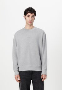 Gray textured sweatshirt with ribbed cuffs and waistband. Small logo on the chest. Model stands in front of a plain white background.
