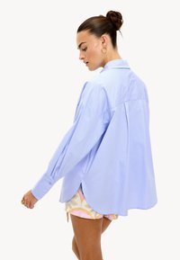 Mujer con el cabello recogido en un moño, vistiendo una camisa de gran tamaño en un azul claro, pantalones cortos con estampados, aretes de aro dorados y un anillo, de pie frente a un fondo blanco.