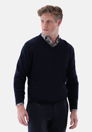  navyfarbener V-Ausschnitt-Pullover aus weichem Stoff, mit langen Ärmeln und einem darunter getragenen Hemd mit Kragen, das ein Blumenmuster zeigt.