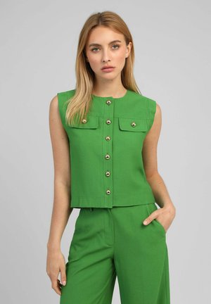 Femme portant un haut vert sans manches à boutons et un pantalon vert assorti, debout la main dans la poche sur un fond gris.
