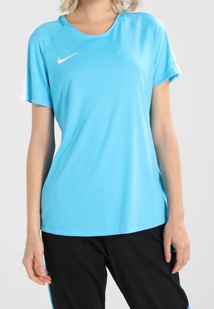 Frau trägt ein hellblaues Nike-Sportshirt mit kurzen Ärmeln und schwarze Hosen mit blauen Streifen, steht vor einem schlichten Hintergrund.