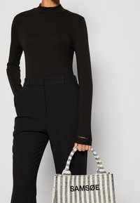 Schwarzes Langarmoberteil, kombiniert mit maßgeschneiderten schwarzen Hosen. Hält eine strukturierte weiße und schwarz gestreifte Tragetasche mit dem Schriftzug "SAM SøE".