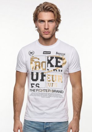 Junger Mann mit blondem Haar, der ein weißes T-Shirt mit gold-schwarzem "Boxeur"-Schriftzug und Logos trägt, steht vor einem schlichten weißen Hintergrund.