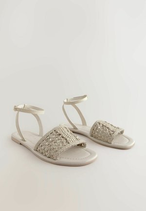 Sandali piatti con tomaia in tessuto beige intrecciato e suola liscia di colore chiaro. Presentano cinturini alla caviglia regolabili per una vestibilità sicura.
