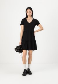 Black tiered dress with short sleeves and a V-neck. Paired with a floral drawstring bag and black chunky shoes, completed with white socks.