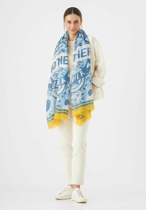 Blauwe en witte sjaal met een patroon, versierd met gele en groene accenten, met florale ontwerpen en tekst, gedrapeerd over een lichte outfit.