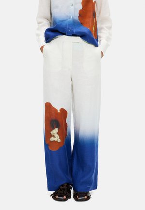 Białe spodnie o szerokich nogawkach z niebieskim gradientem na dole i dużymi brązowymi abstrakcyjnymi plamami, w połączeniu z czarnymi sandałami z odkrytymi palcami.