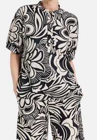 Chemisier à boutons noir et crème à manches courtes, présentant un motif floral. Confectionné en tissu léger avec une coupe décontractée.