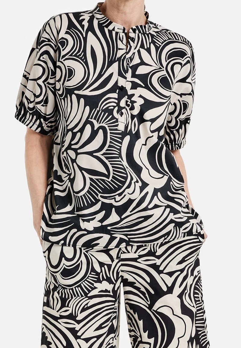 Chemisier à boutons noir et crème à manches courtes, présentant un motif floral. Confectionné en tissu léger avec une coupe décontractée.