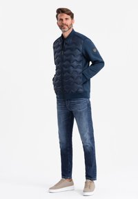 Veste rembourrée bleu marine avec texture ondulée, fermeture éclair et col haut. Associée à un jean bleu et des baskets beige clair.