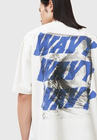 Camiseta de algodón blanca de corte oversize, que presenta un gran texto gráfico azul "WAVY" y un patrón abstracto de olas grises en la parte posterior.