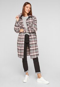 Manteau à carreaux en gris, bordeaux et blanc, boutonné, avec deux poches avant, porté par-dessus un pull gris et un pantalon noir, associé à des baskets blanches.
