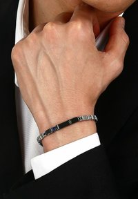 Silberarmband mit schwarzen Akzenten, ausgestattet mit einer glatten rechteckigen Mitte und blauen Edelstein-Details. Am nackten Handgelenk getragen.