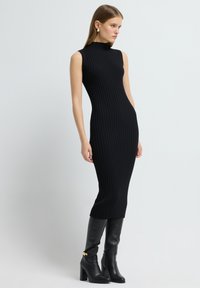 Czarna dzianinowa sukienka prążkowana z wysokim kołnierzem i bezrękawowym krojem, zestawiona z czarnymi skórzanymi botkami do kolan z złotymi akcentami.