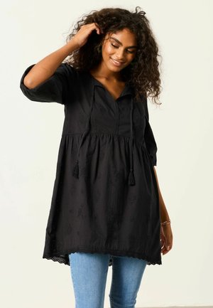 Femeie cu păr creț purtând o tunică largă neagră brodată și blugi albaștri, zâmbind și atingându-și părul cu o mână.