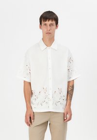 Hvid kortærmet skjorte med blomstret lace-detaljer ved kanten og ærmerne, med knaplukning foran og spidse klapper.