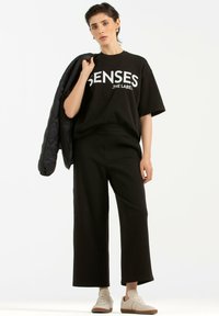 Schwarzes oversized T-Shirt mit weißen grafischen Text, schwarze weite Hosen und beige Sneaker. Eine gesteppt schwarze Jacke hängt über einer Schulter.