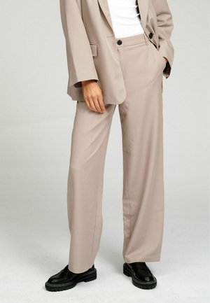 Személy bézs bő szárú nadrágban, hozzáillő blézerben, fehér felsőben és fekete masszív loaferekben, egyik kezét zsebében tartva áll.