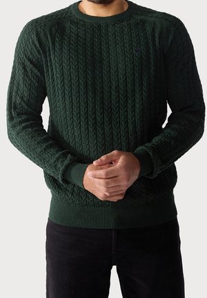 Mann trägt einen dunkelgrünen Pullover mit Zopfmuster, langen Ärmeln und gerippten Bündchen, kombiniert mit schwarzen Hosen, Hände vor dem Körper verschränkt.