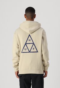 Šviesiai rudas hoodie su dideliu jūros mėlynumo trikampio grafiniu raštu ant nugaros, kuriame yra raidės H, U ir F. Minkšta medžiaga, laisvas siluetas.