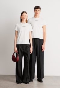 Λευκά t-shirts με το λογότυπο "TOMMY", σε συνδυασμό με μαύρο παντελόνι φαρδιάς γραμμής. Η γυναίκα κρατάει μια μπορντό τσάντα με καμπυλωτό σχήμα. Μινιμαλιστικός σχεδιασμός.