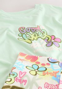 Chemise vert clair avec texte coloré « Good Vibes » et motif fleur, associée à un tissu à motifs floraux multicolores et style graffiti.