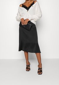 Krämfärgad ribbad cardigan med stora knappar, svart satin klänning, svart kuvertväska och svarta spänna klacksandaler. Lätta texturer och eleganta linjer.