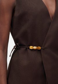 Vêtement sans manches brun foncé avec trois perles décoratives marron clair sur une ceinture à nouer sur le côté à la taille.