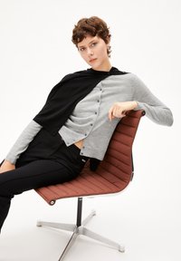 Grijze en zwarte button-up cardigan gedragen over een grijze top, gecombineerd met een zwarte broek, zittend op een moderne, gevoerde, bruine stoel met een zilveren basis.