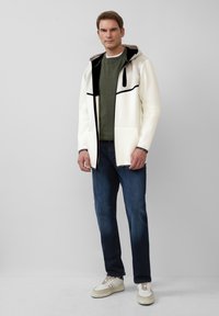 Weißer Reißverschluss-Jacke mit einer Kapuze in Schwarz und Grau-Muster, getragen über einem dunkelgrünen Shirt und kombiniert mit dunklen Jeans und weißen Turnschuhen.