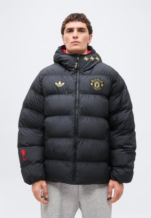 Hombre con chaqueta acolchada negra de Manchester United con logotipos dorados y emblema rojo en la manga, de pie frente a un fondo liso.