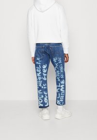 Vaqueros de mezclilla azul desgastados con texto pintado en blanco, corte recto y costuras en contraste. Combinados con una sudadera blanca con capucha y zapatillas.