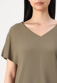 Top vert olive à col en V en tissu lisse, présentant des manches courtes et une coupe décontractée. L'encolure est modestement découpée pour plus de confort.
