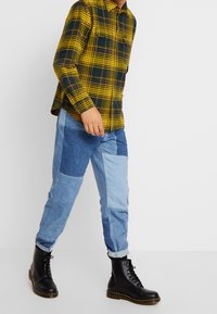 Geel-groen geruit overhemd met twee borstzakken, gecombineerd met patchwork blauwe jeans en zwarte veterlaarzen.