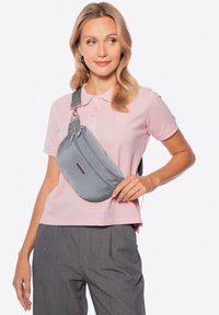 Šedá látková křížová taška s dvěma zipovými přihrádkami, nošená přes tělo, v kombinaci se světle růžovým polo tričkem a šedými kalhotami.