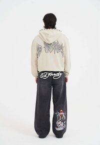 Felpa beige con logo grigio e cappuccio regolabile. Pantaloni in denim neri con logo bianco e grafica colorata sulla tasca posteriore.