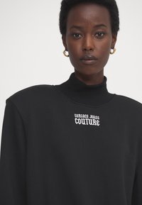 Sudadera negra con un cuello alto, que presenta bordado blanco de "VERSACE JEANS COUTURE" en el pecho. Textura suave, diseño minimalista.