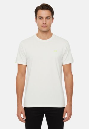 Uomo con capelli castani corti che indossa una maglietta bianca a maniche corte semplice e pantaloni neri, in piedi davanti a uno sfondo bianco.